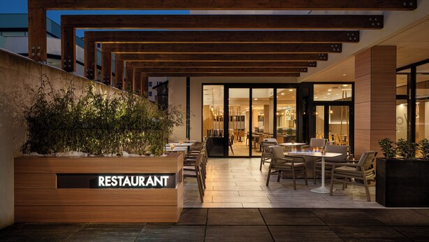 Un espace extérieur aménagé pour les repas, avec des pergolas en bois, une enseigne indiquant « RESTAURANT » et une vue sur l’intérieur du bâtiment.