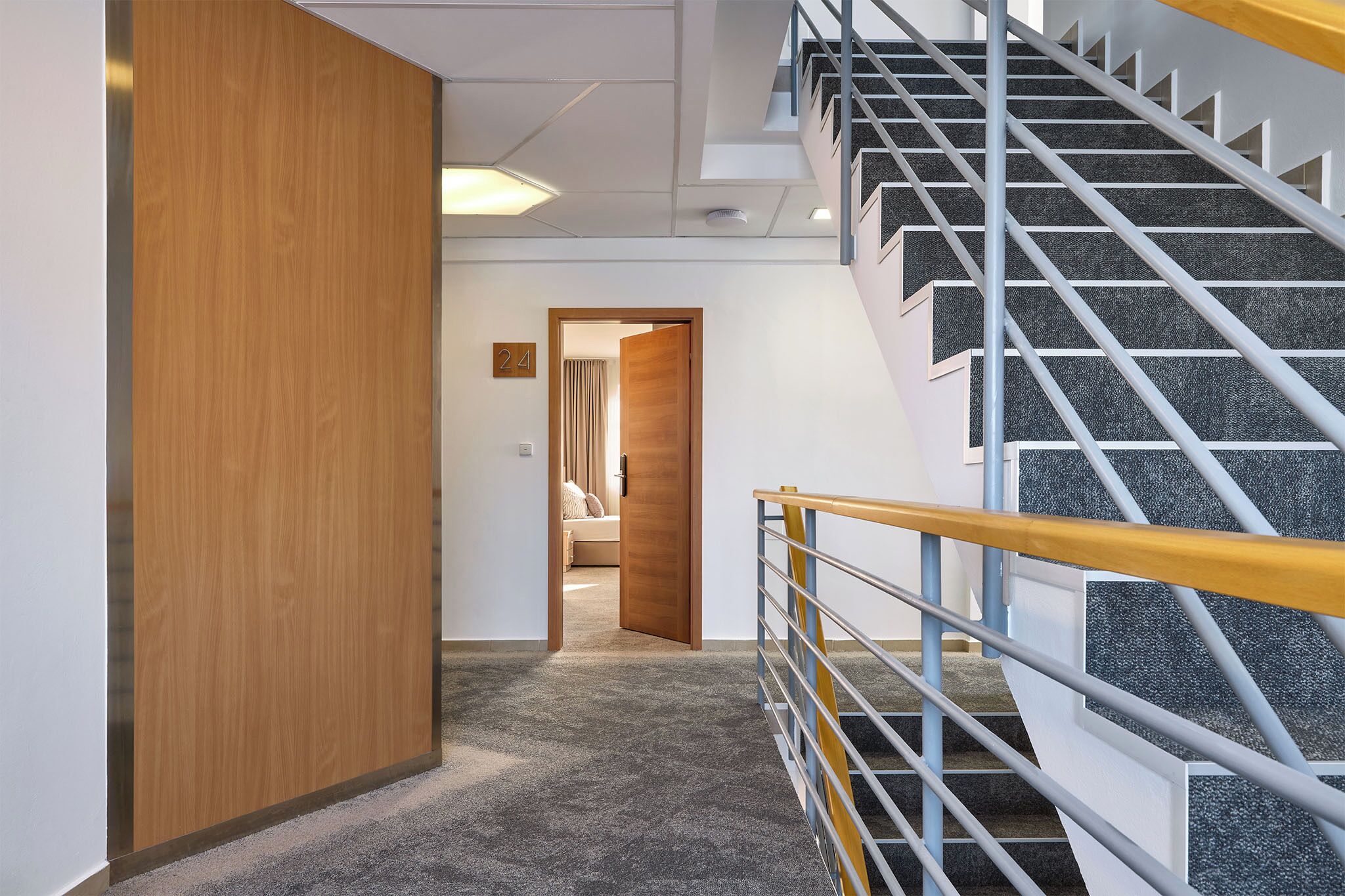 Un interior moderno con pasillo alfombrado, puerta de madera y escalera con barandilla de metal.