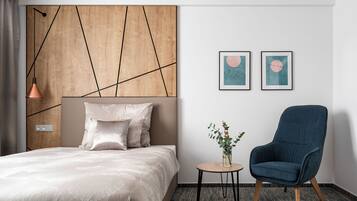 A hotel room with a bed, a blue armchair, a small table with a vase, and two framed artworks on the wall.