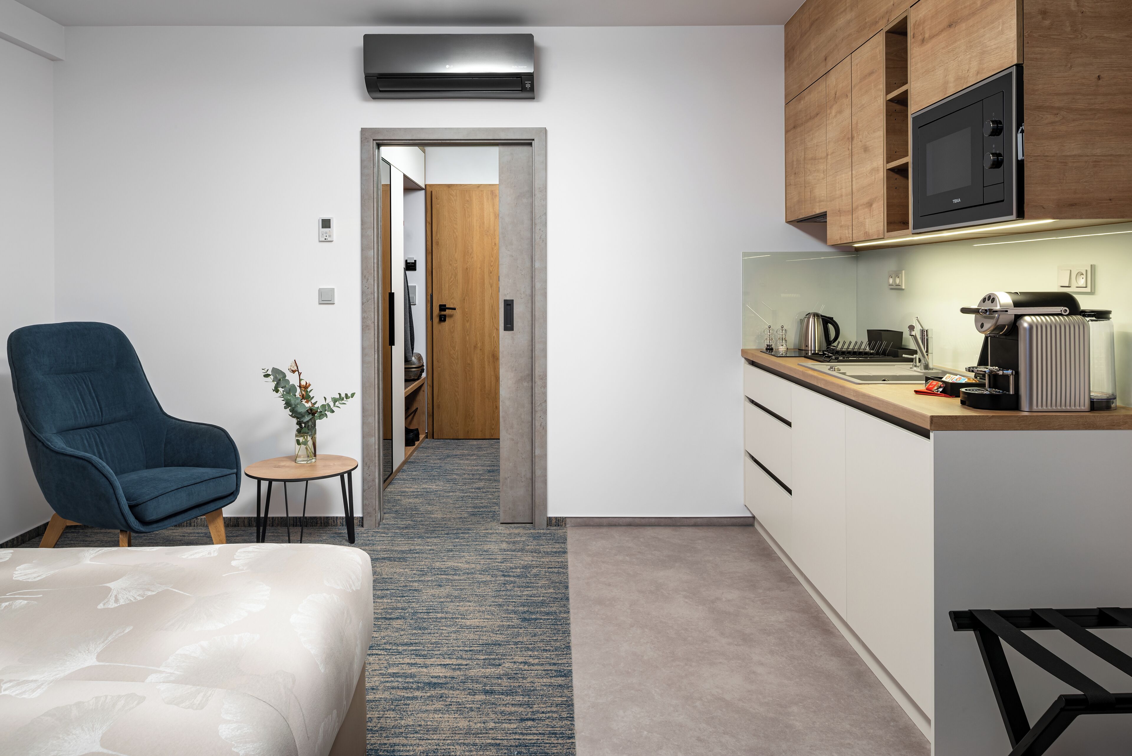 Una habitación de hotel moderna con una cama, un sillón azul, una mesita con un jarrón, una kitchenette con microondas y una puerta de madera que da a otra habitación.