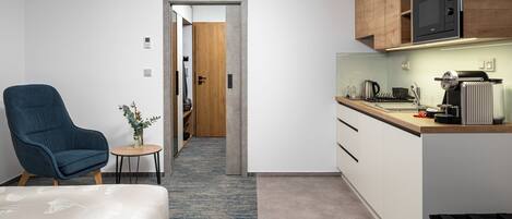 Una habitación de hotel moderna con una cama, un sillón azul, una mesita con un jarrón, una kitchenette con microondas y una puerta de madera que da a otra habitación.
