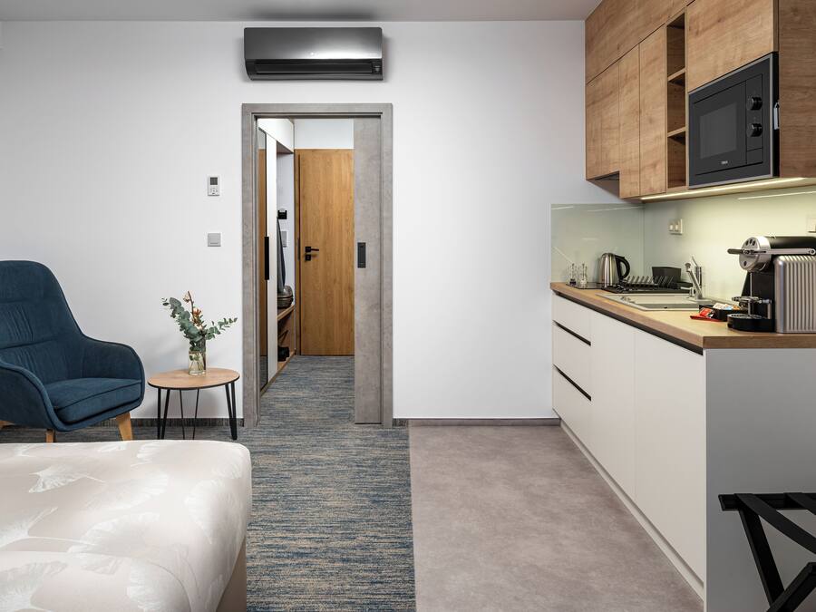 A modern hotel room with a bed, a blue armchair, a small table with a vase, a kitchenette with a microwave, and a wooden door leading to another room.