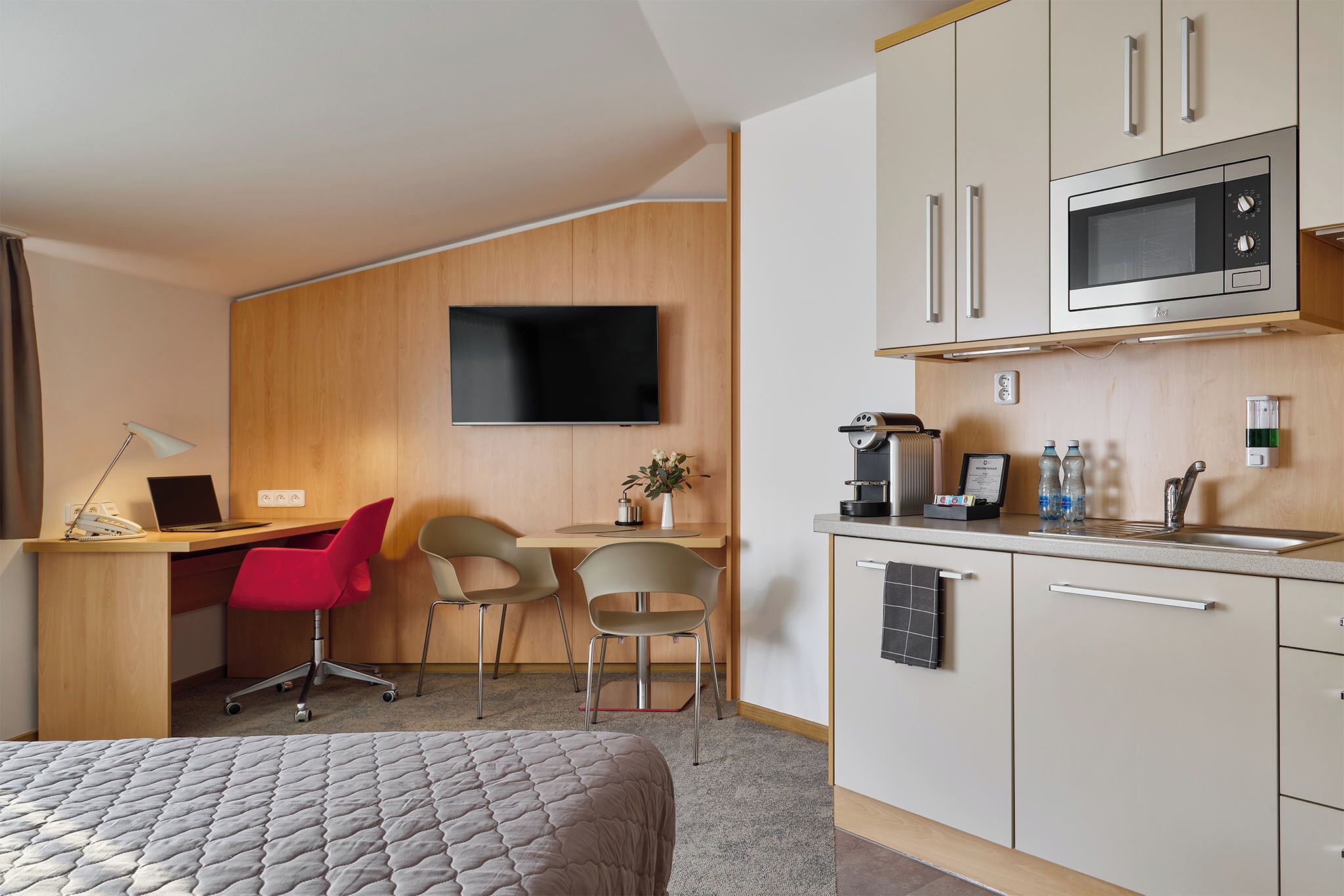 Une chambre d’hôtel comprenant un lit, un bureau avec un ordinateur, une télévision, une kitchenette équipée d’un four à micro-ondes et un petit coin repas.