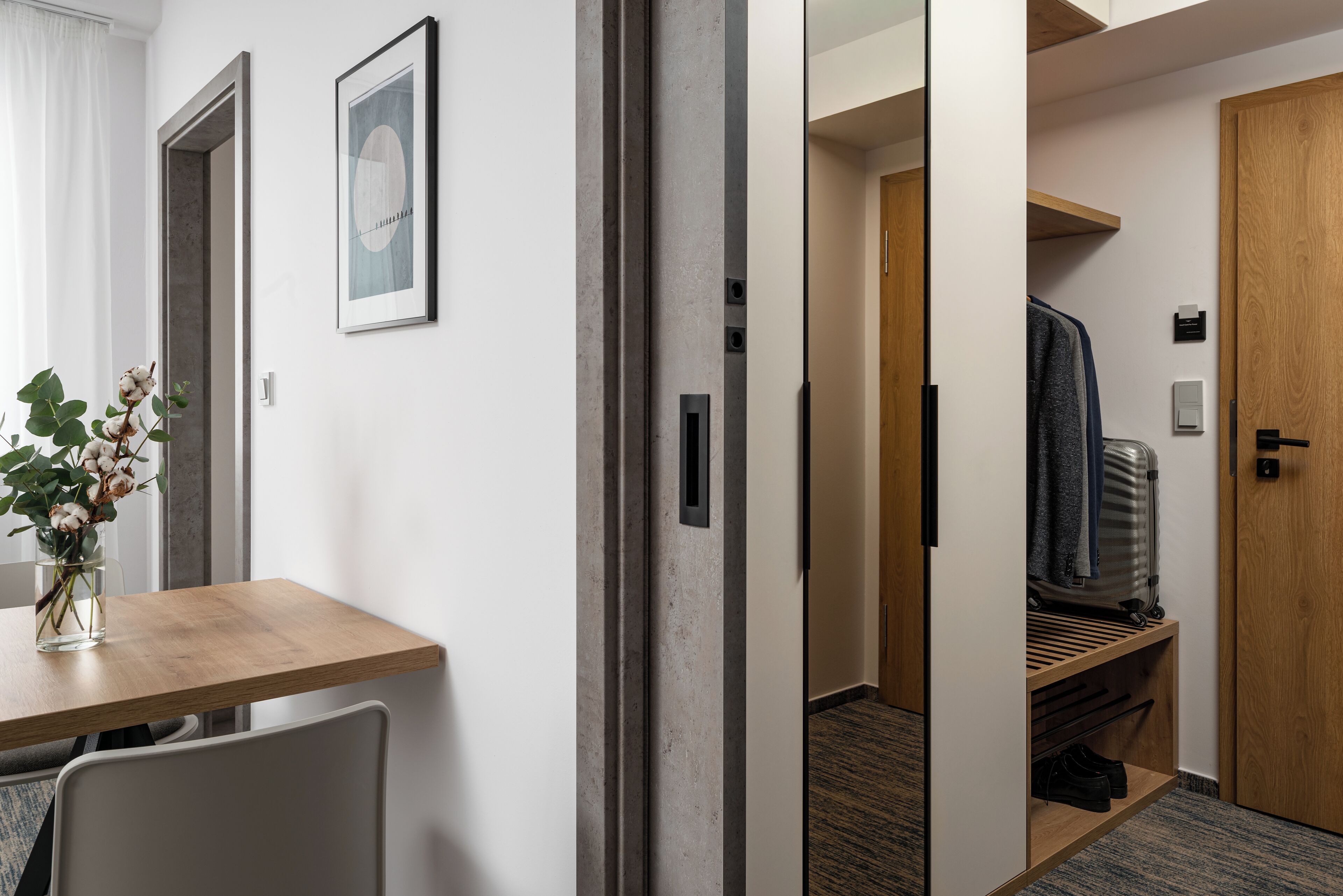 Habitación de hotel con escritorio de madera, un florero con flores, un cuadro en la pared y un armario con ropa y zapatos colgados.