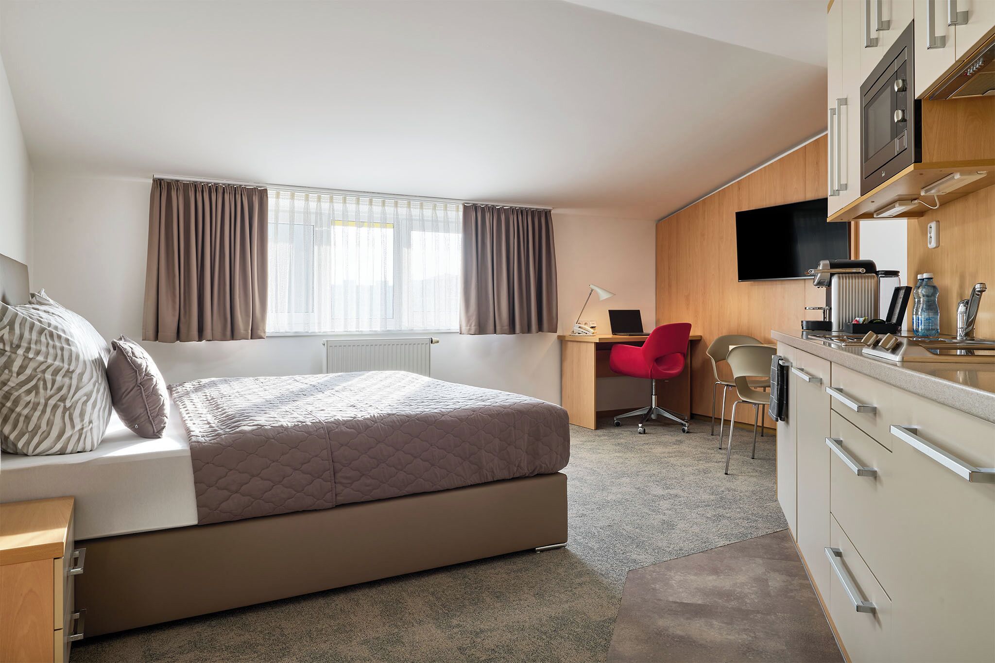 A compact hotel room with a bed, a desk, a chair, a TV, and a kitchenette.