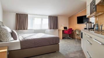 A compact hotel room with a bed, a desk, a chair, a TV, and a kitchenette.
