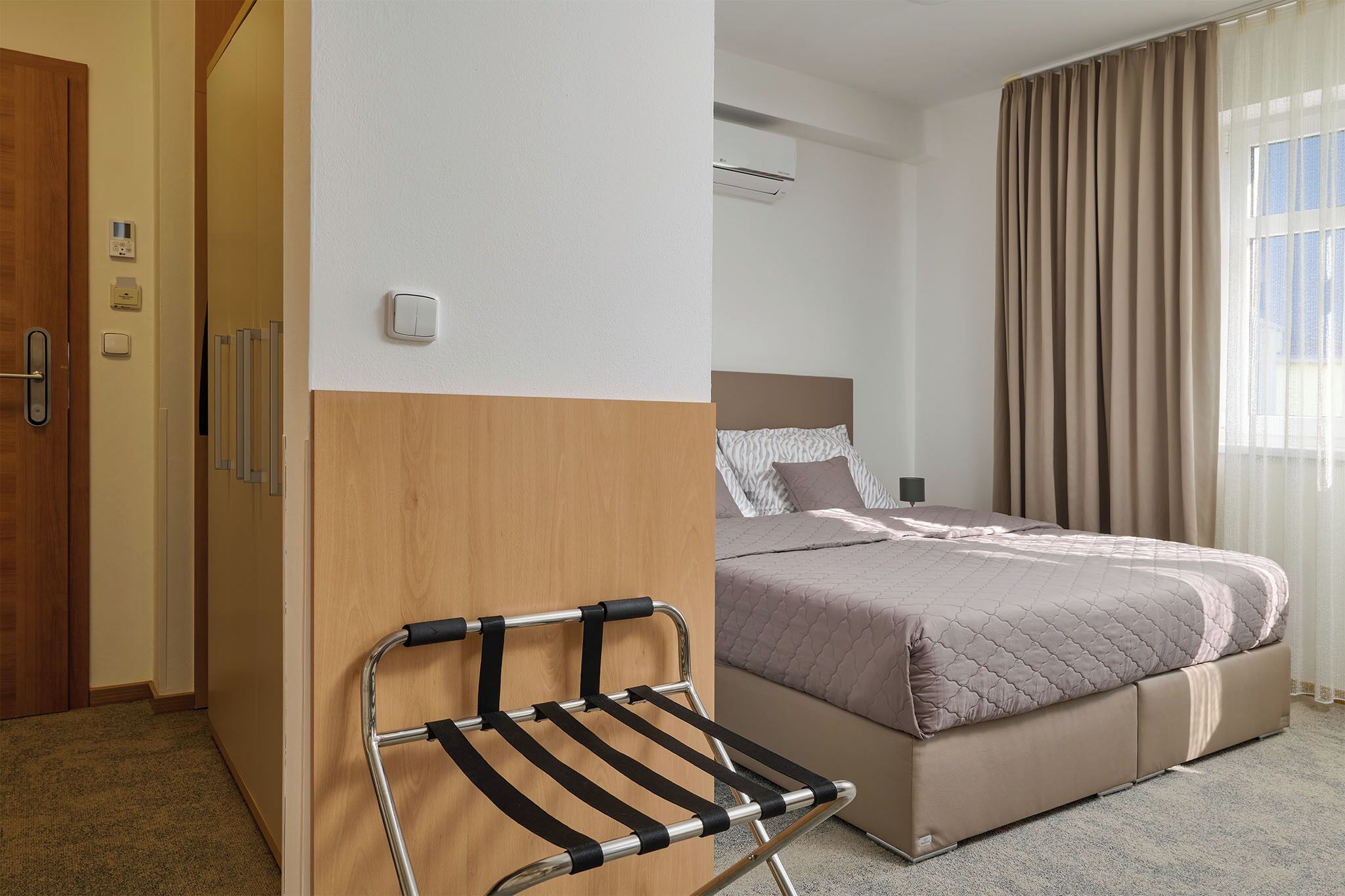 A hotel room with a bed, a chair, a wooden wardrobe, and a door.