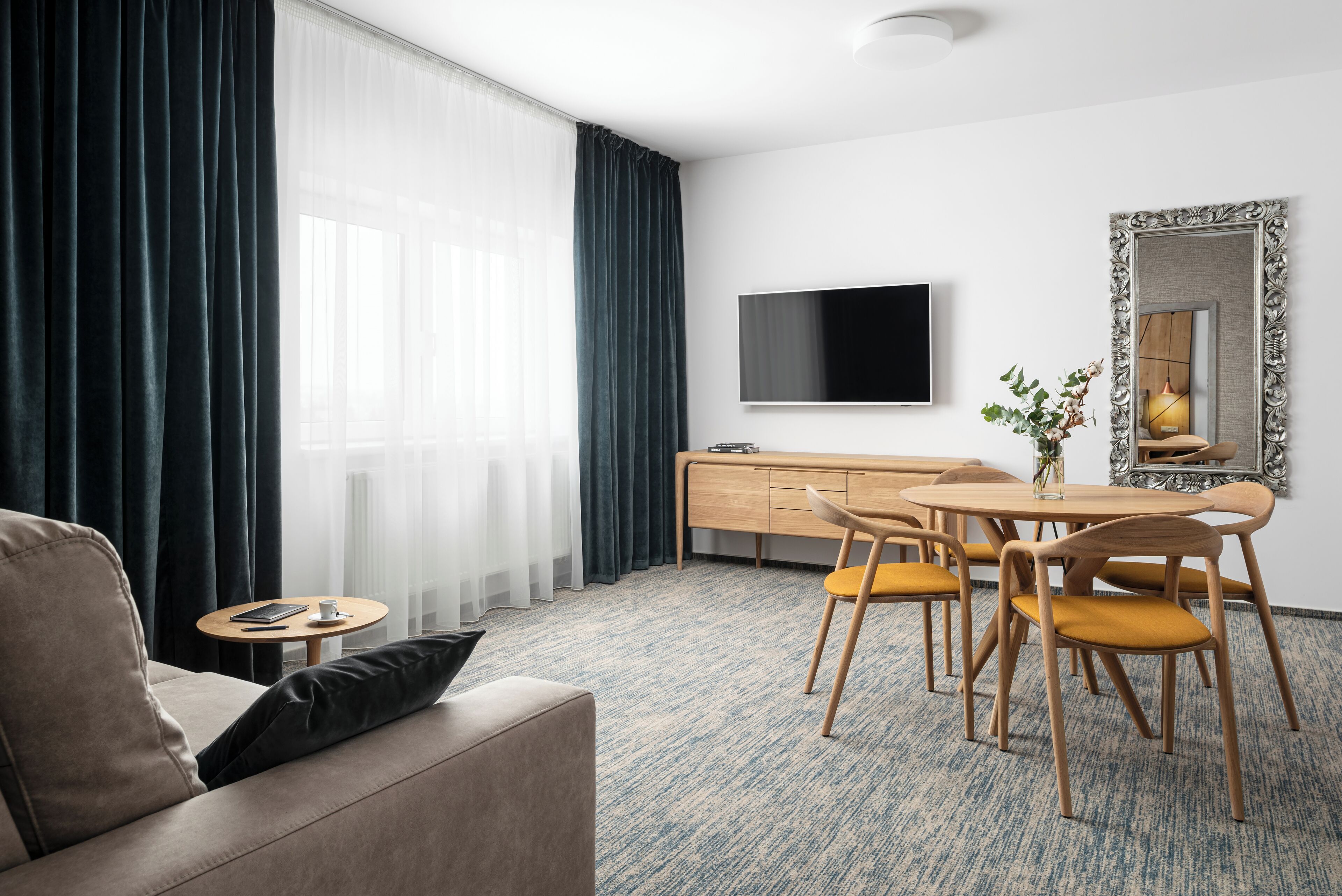 A modern hotel room with a sofa, a dining table with chairs, a TV mounted on the wall, and a large mirror.