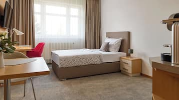 Quarto de hotel com cama, escrivaninha, cadeira, televisão e mesa de cabeceira.