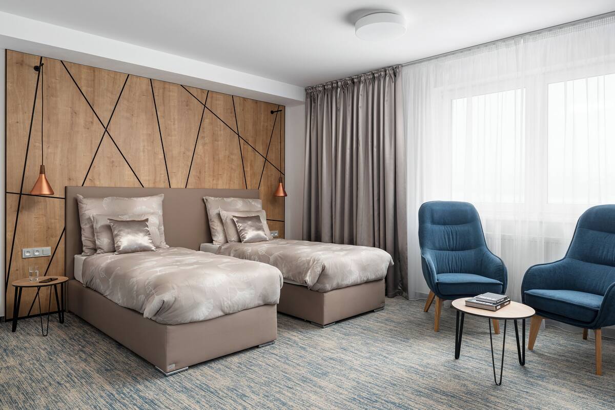 Une chambre d’hôtel avec deux lits, une petite table, deux chaises, un mur décoratif en bois et une grande fenêtre avec des rideaux.