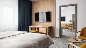 Une chambre d’hôtel dotée d’un grand lit, d’un bureau avec une chaise, d’un téléviseur fixé au mur et d’un coin repas visible à travers une porte ouverte.
