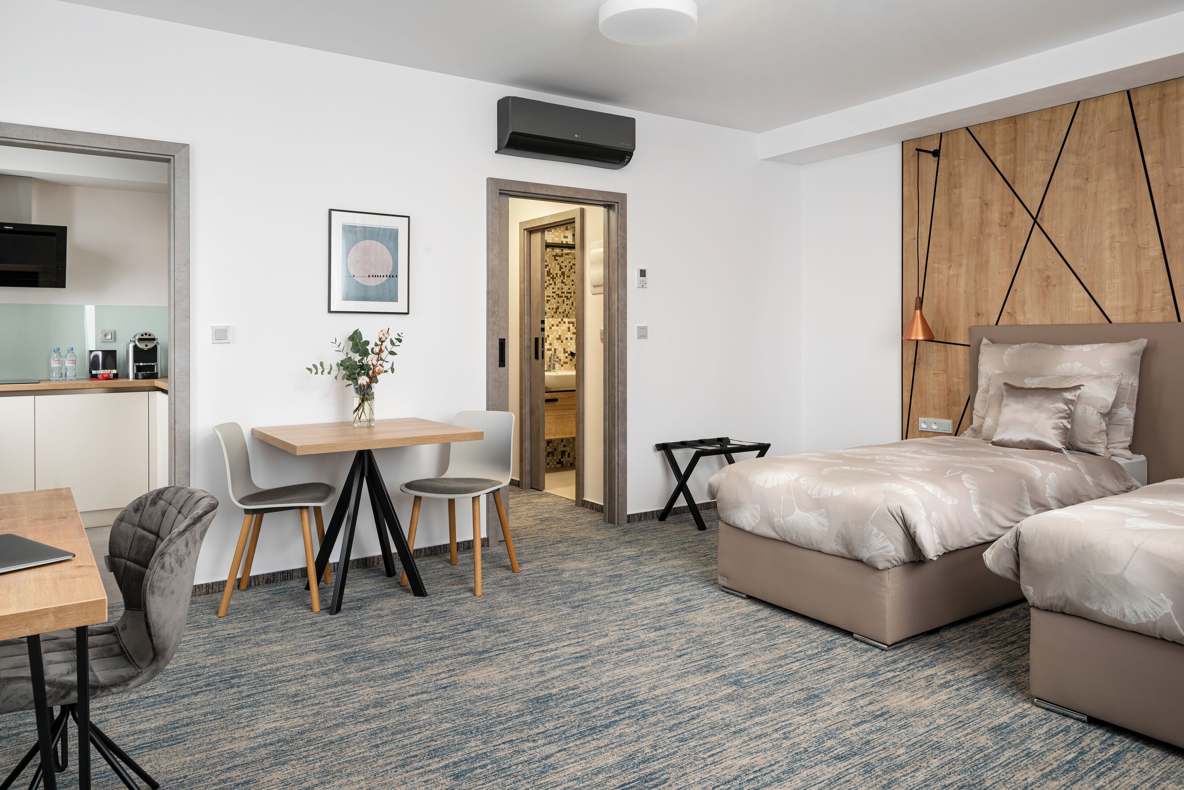 Moderni hotellihuone, jossa on sänky, työpöytä ja tuoli. Huoneessa on pieni pöytä, jonka päällä on maljakko täynnä kukkia. Huoneessa on puinen sängynpääty ja seinään kiinnitetty ilmastointilaite.