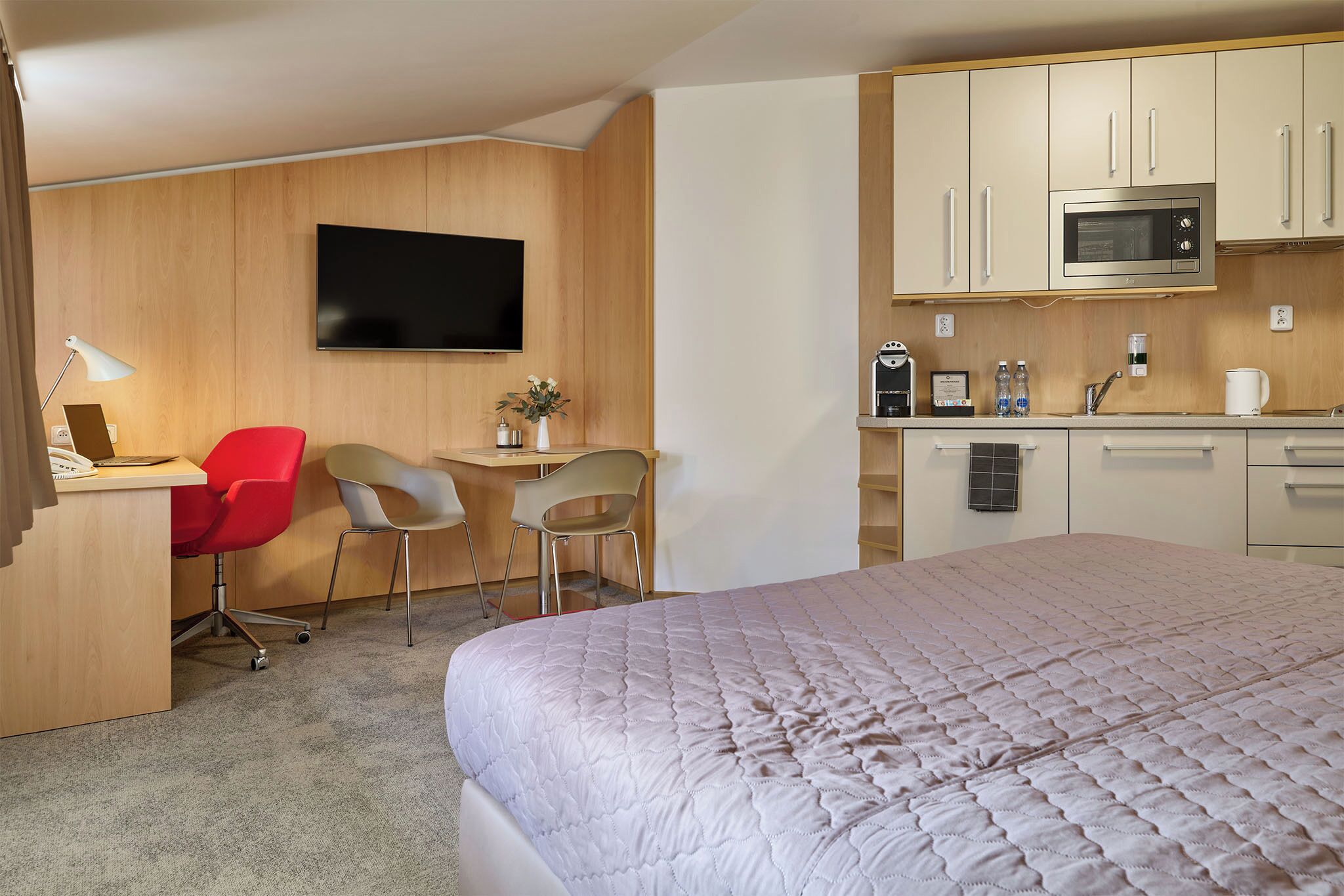A hotel room with a bed, a desk with two chairs, a television, a microwave, and a small kitchenette.