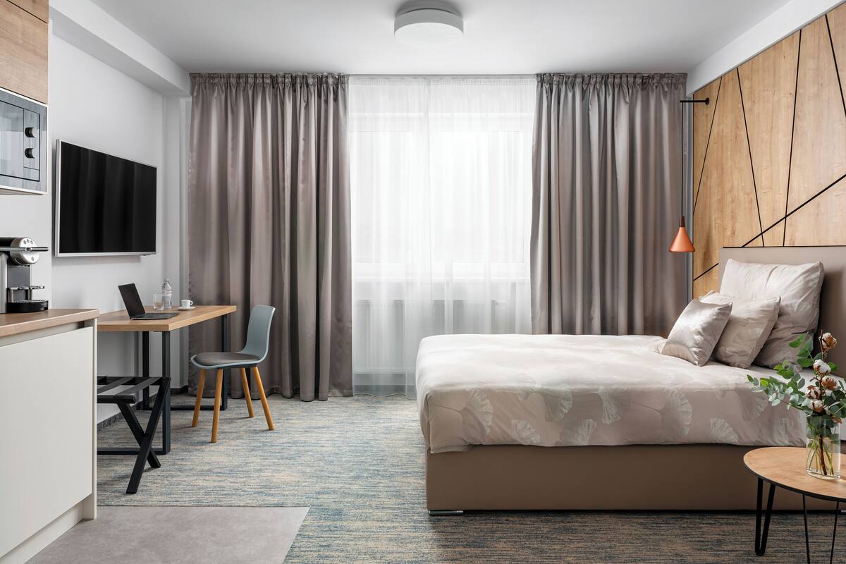 Une chambre d’hôtel moderne équipée d’un lit, d’un bureau avec un ordinateur portable, d’une chaise, d’une télévision et d’une grande fenêtre avec des rideaux.