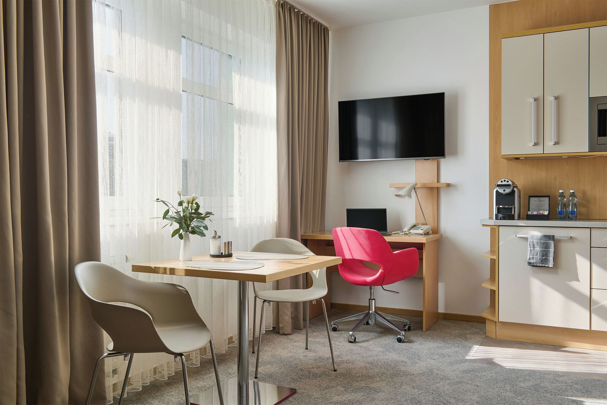 A hotel room with a desk, two chairs, a TV, a minibar, and a large window with curtains.