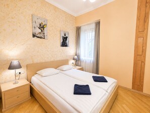 A hotel room with a double bed, two bedside tables, a nightstand, a lamp, a window with curtains, and artwork on the wall. - Charming Old Town Apartment next to the Iconic Swedish Gate (Riga)