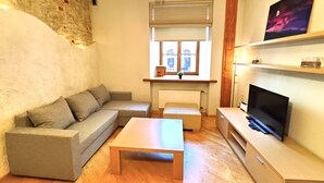 Een woonkamer met een grijze bank, een houten salontafel, een tv op een houten dressoir, en een raam met uitzicht op gebouwen.