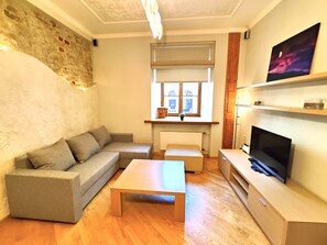 A living room with a grey sofa, a wooden coffee table, a TV on a wooden stand, and a window with a view of buildings. - Swedish Gate One Bedroom Apartments Old Town Riga (Riga)