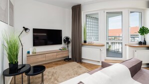Ein modernes Wohnzimmer mit einem Flachbildfernseher, einem Standventilator, einem kleinen Tisch mit einer Pflanze, einem Sofa und einem großen Fenster mit Blick auf Gebäude.