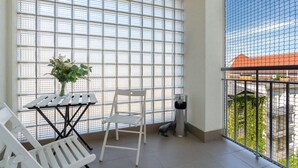 Ein Balkon mit einem weißen Stuhl, einem kleinen Tisch mit Blumen in einer Vase und einer Metallmülltonne.