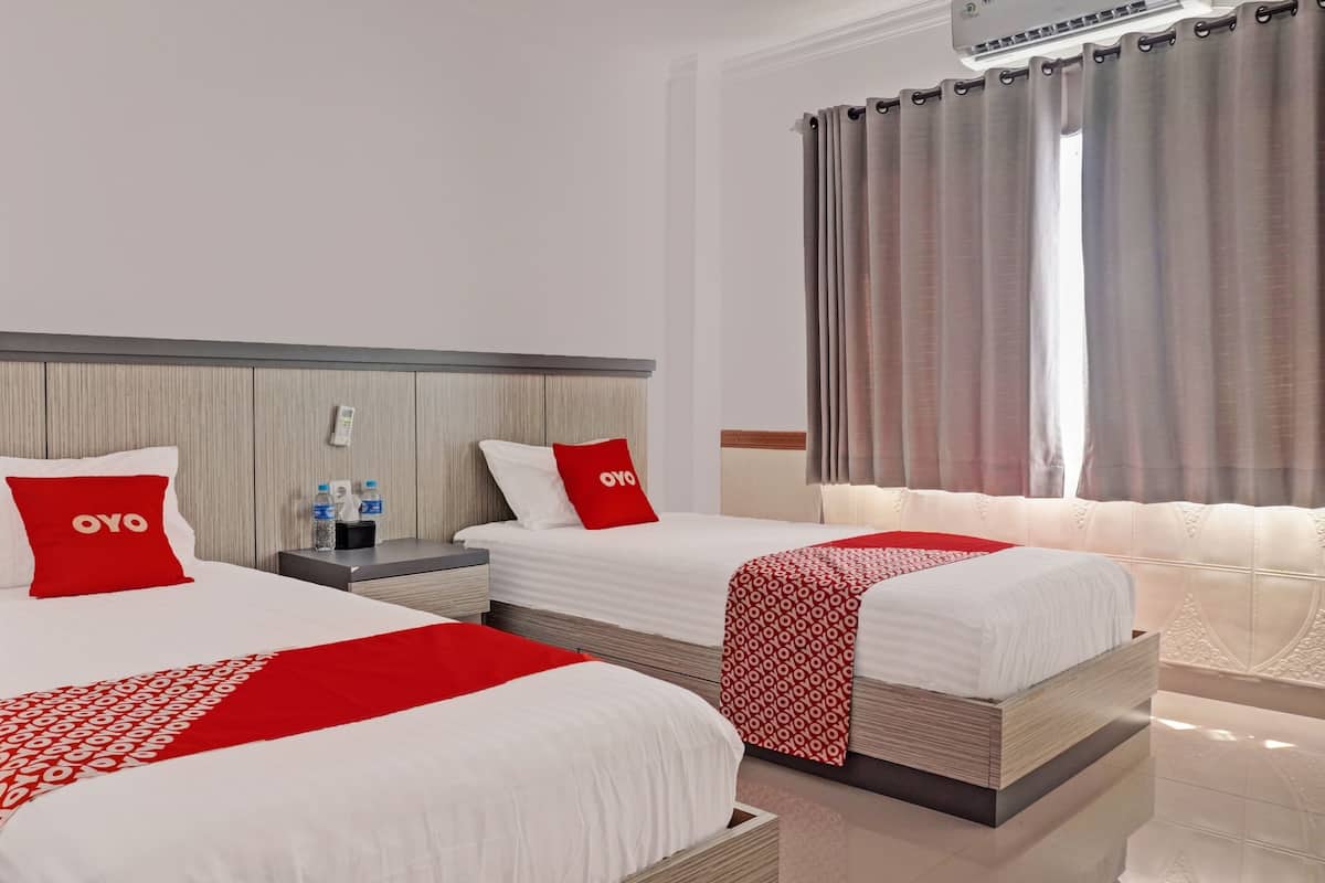A hotel room with two beds, each with a white bedspread and red pillows. There is a nightstand with a water bottle and a phone. The room has a large window with curtains.