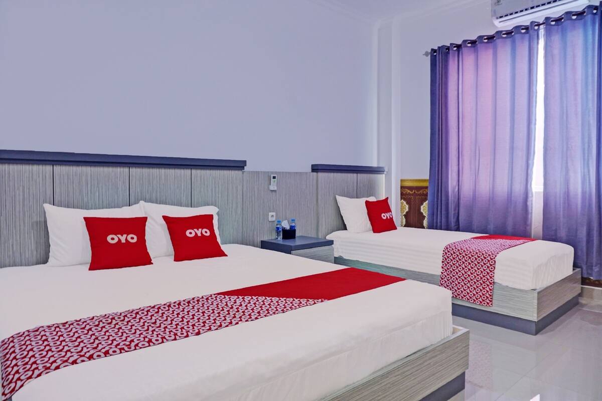 A hotel room with two beds, each with a red and white patterned bedspread, and matching red pillows. There is a nightstand with a water bottle and a phone charger. The room has a large window with purple curtains.