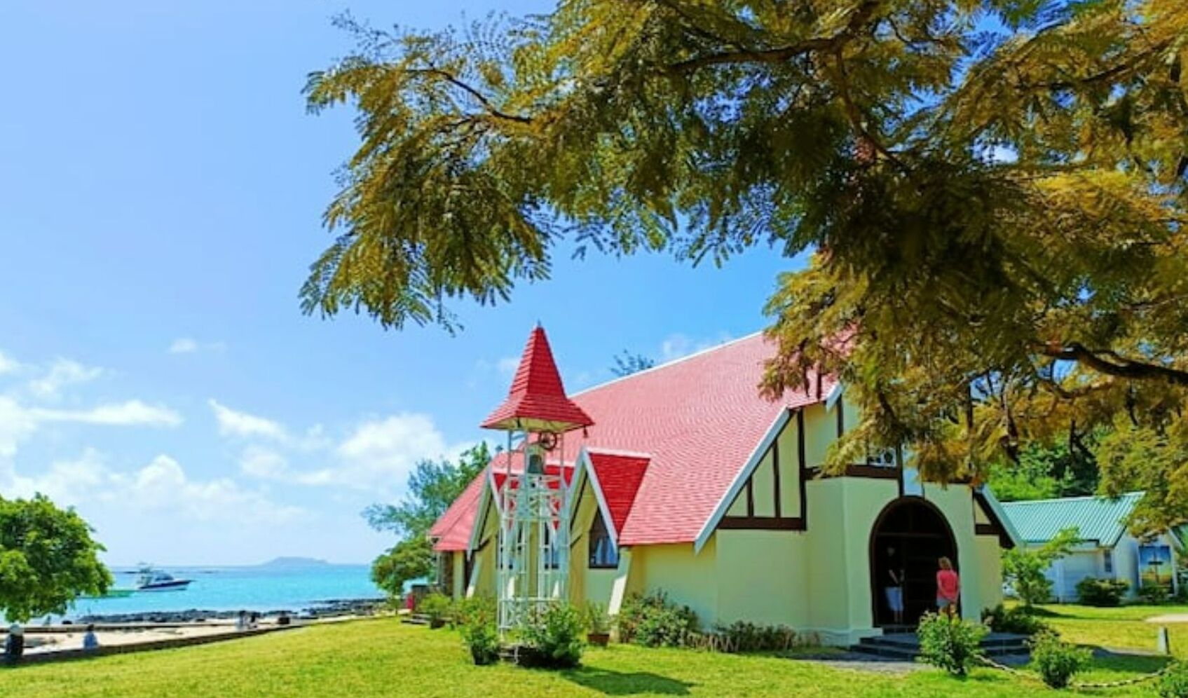 A church with a red roof and a bell tower, surrounded by greenery and a clear blue sky.