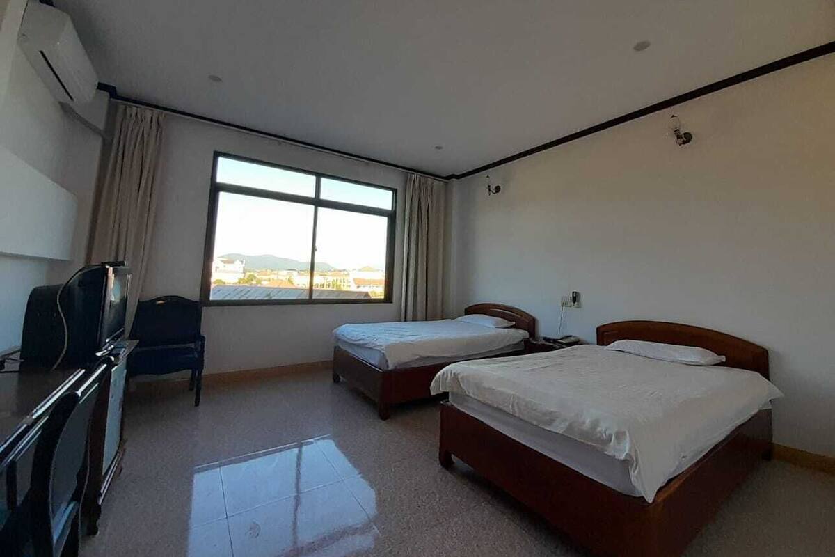 Ein Hotelzimmer mit zwei Einzelbetten, einem Schreibtisch mit Stuhl, einem Fernseher und einem Fenster mit Vorhängen.