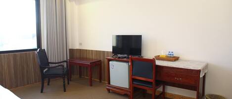 A hotel room with a desk, two chairs, a television, a small refrigerator, and a window with curtains.