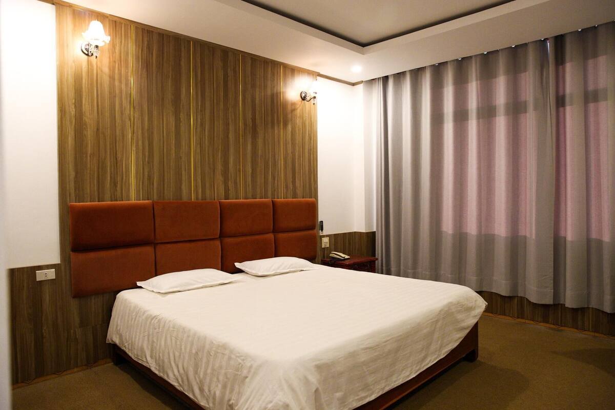 Ein Hotelzimmer mit einem Holzkopfteil, einem großen Bett mit weißen Leinen, einem Nachttisch mit einer Lampe und Blick durch Vorhänge nach draußen.