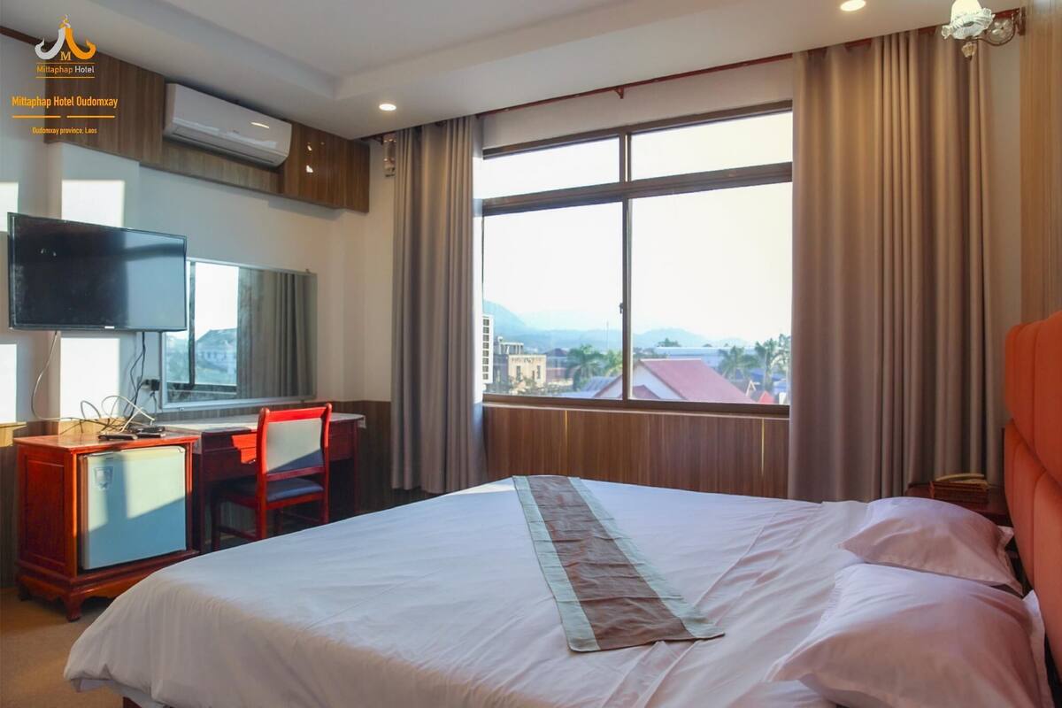 Ein Hotelzimmer mit einem Bett, einem Schreibtisch mit Stuhl, einem Fernseher und einem großen Fenster mit Vorhängen.