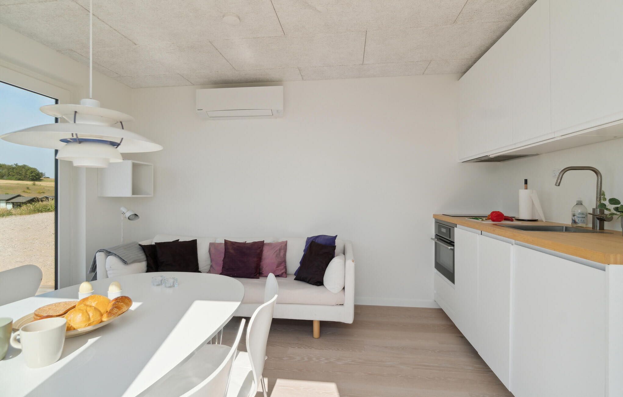 A modern kitchen with white cabinets, a wooden countertop, and a built-in oven. There is a white dining table with a plate of bread and a cup. A white sofa with colorful pillows is in the background.