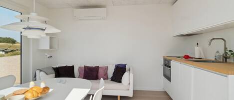 Eine moderne Küche mit weißen Schränken, einer Holzarbeitsplatte und einem eingebauten Ofen. Es gibt einen weißen Esstisch mit Brot und einer Tasse. Im Hintergrund steht ein weißes Sofa mit bunten Kissen.