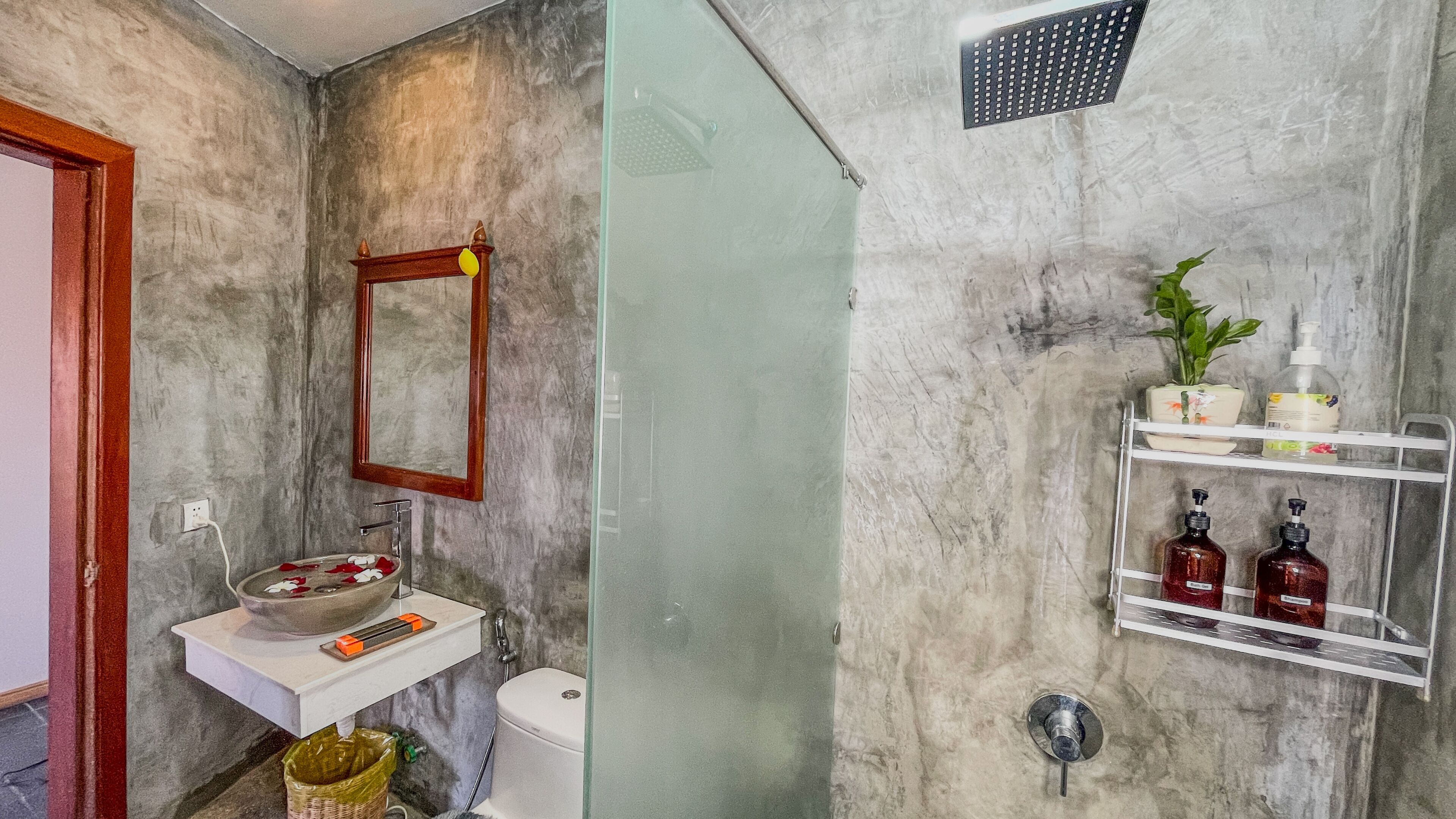 A bathroom with a marble wall, a wooden-framed mirror, a white sink with a bowl, a toilet, and a shower area with a glass door. There are shelves with toiletries and a potted plant.