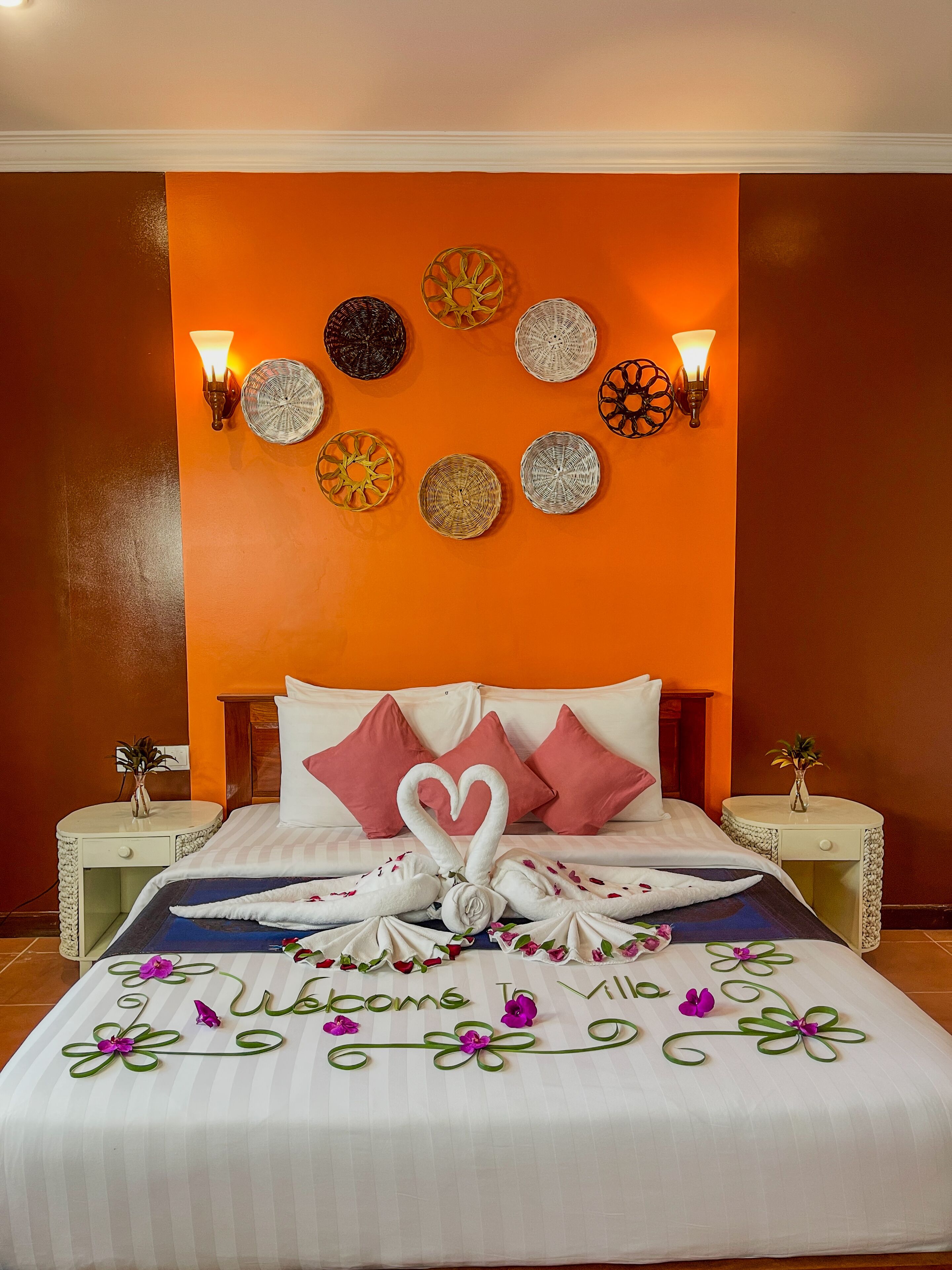 A hotel room with an orange accent wall, a bed with white and pink pillows, and a heart-shaped towel arrangement.
