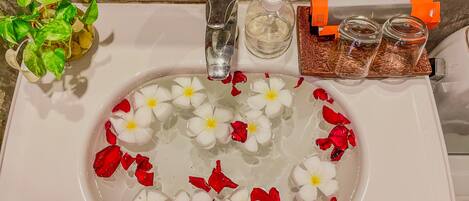 A bathroom sink with water, white flowers, and red petals. There is a plant, a soap dispenser, and a container with a bottle and a box.