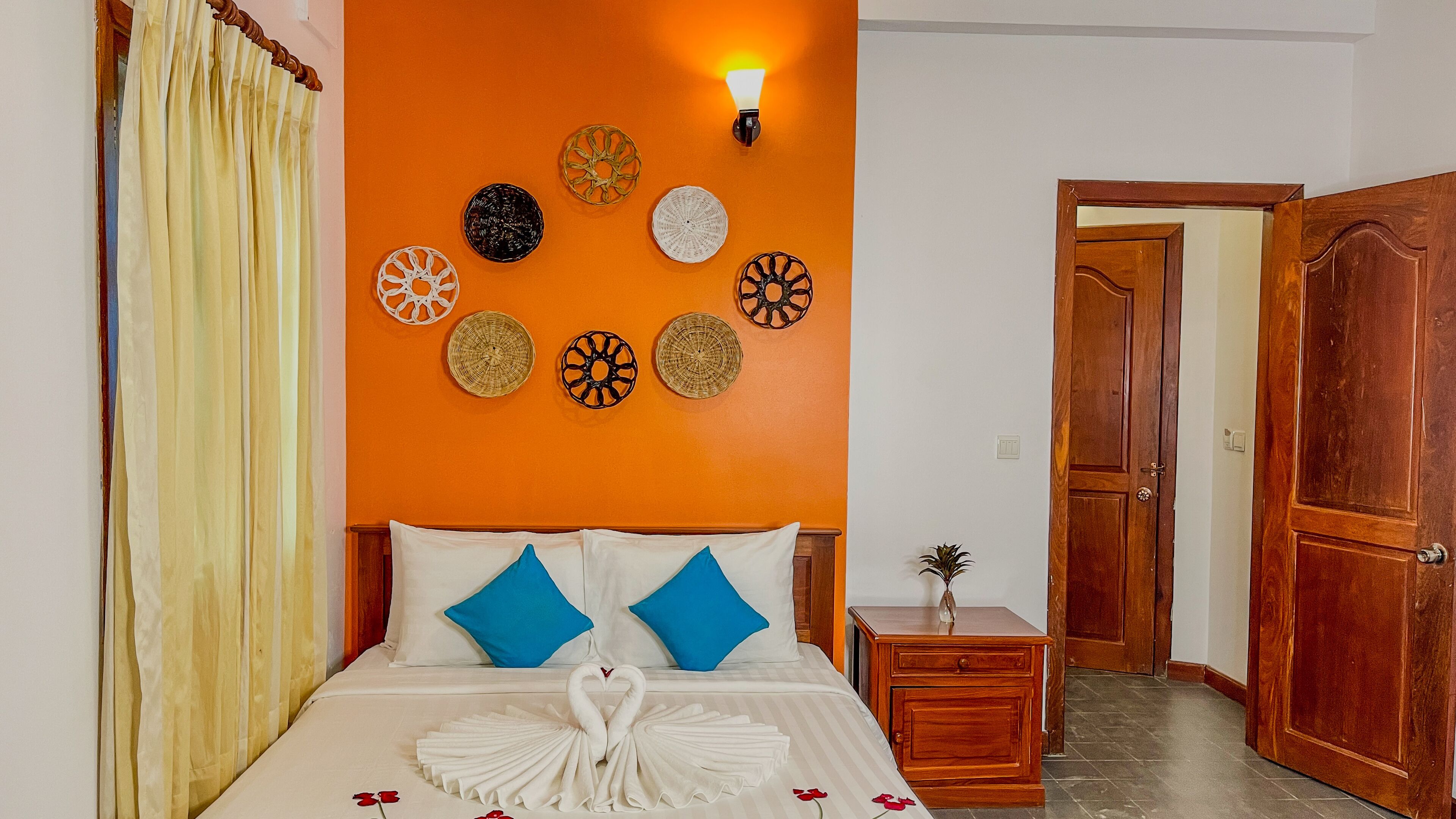 Une chambre d’hôtel avec un mur d’accent orange, un lit avec une literie blanche et bleue, une table de chevet avec une lampe et une porte en bois.