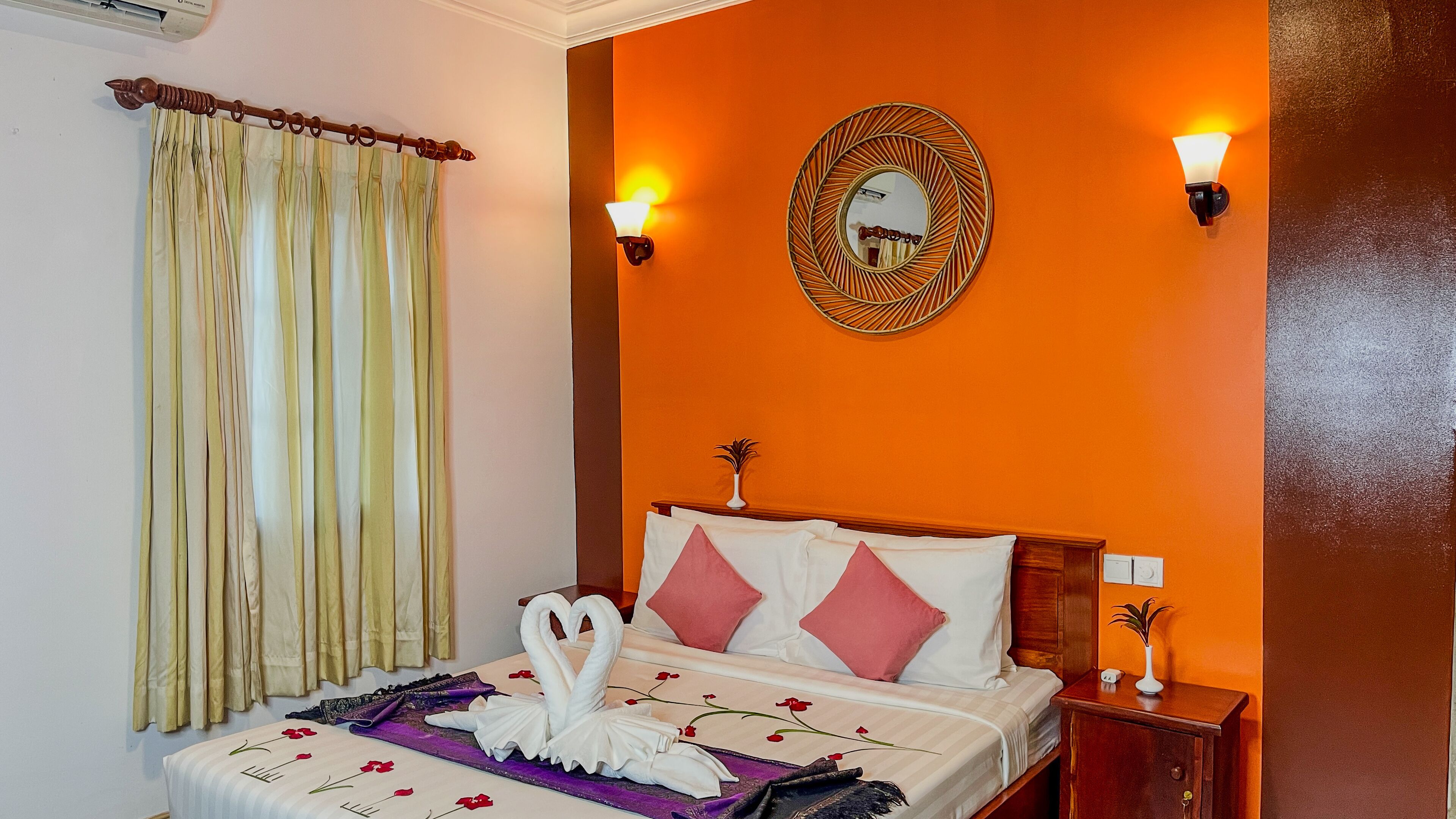 A hotel room with an orange wall, a bed with white and pink pillows, a decorative mirror, and two wall-mounted lights.