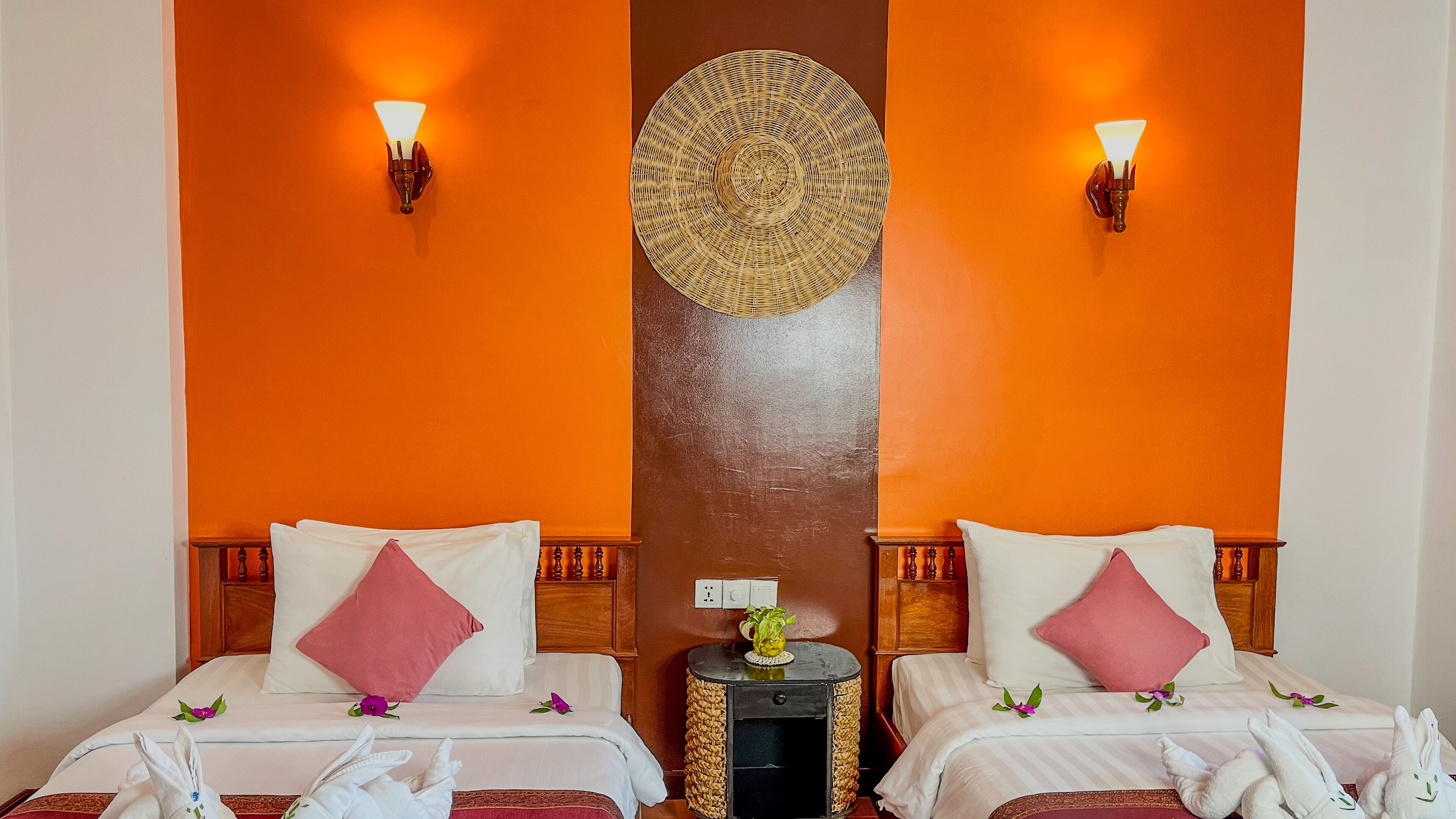A hotel room with two beds, each with white linens and pink pillows, a small round side table with a woven basket, and two wall-mounted lamps.
