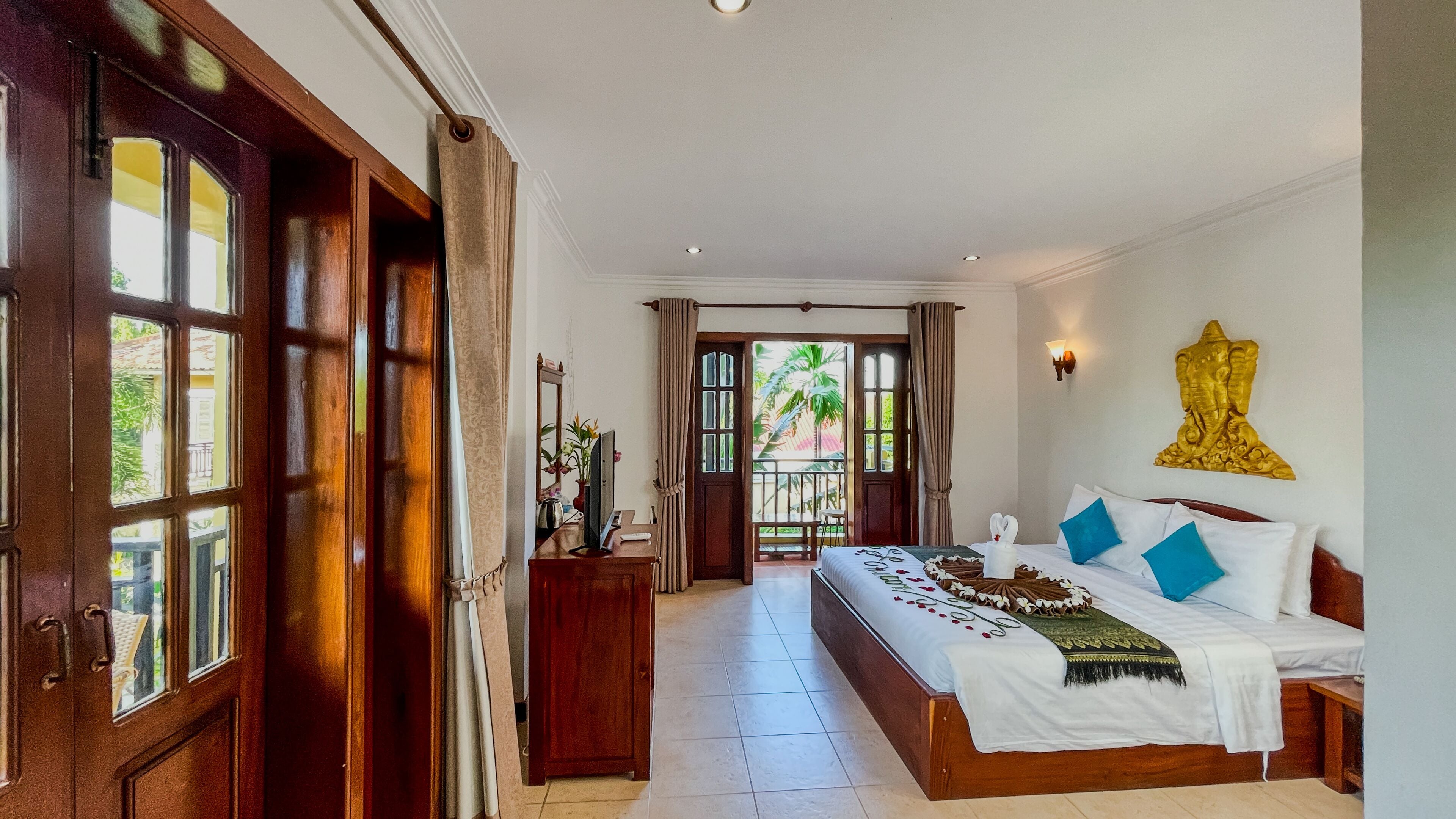 Een hotelkamer met een groot bed, een houten deur, een balkon met uitzicht op palmbomen en een gouden beeldhouwkunst aan de muur.