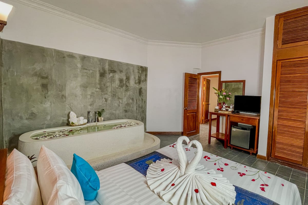 A hotel room with a bathtub, a bed with a decorative swan towel, a desk with a television, and wooden doors.