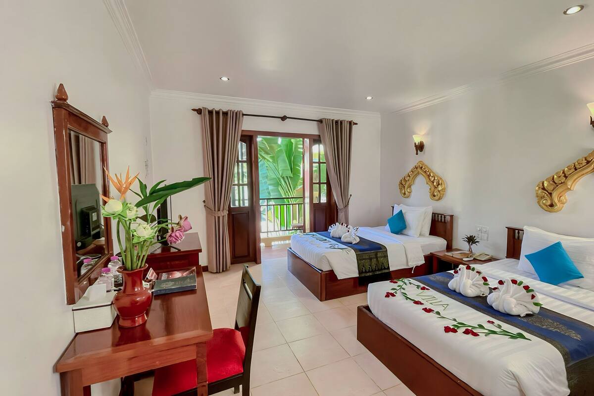 A hotel room with two beds, a desk, and a chair. There is a large window with curtains, a decorative wall piece, and a small table with a vase of flowers.