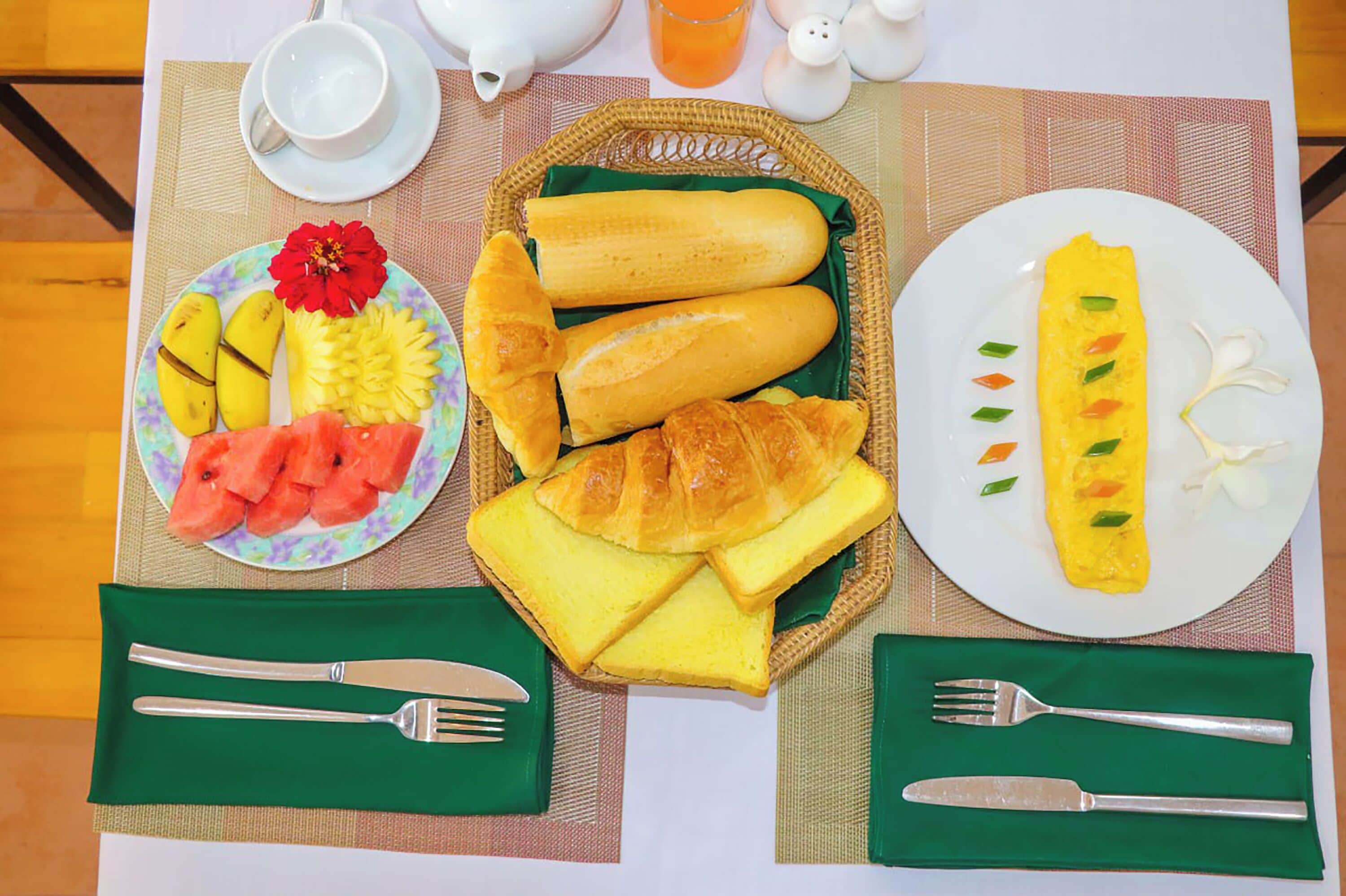 Desayuno con cruasanes, pan, una tortilla y fruta en platos, junto con cubiertos y una tetera.