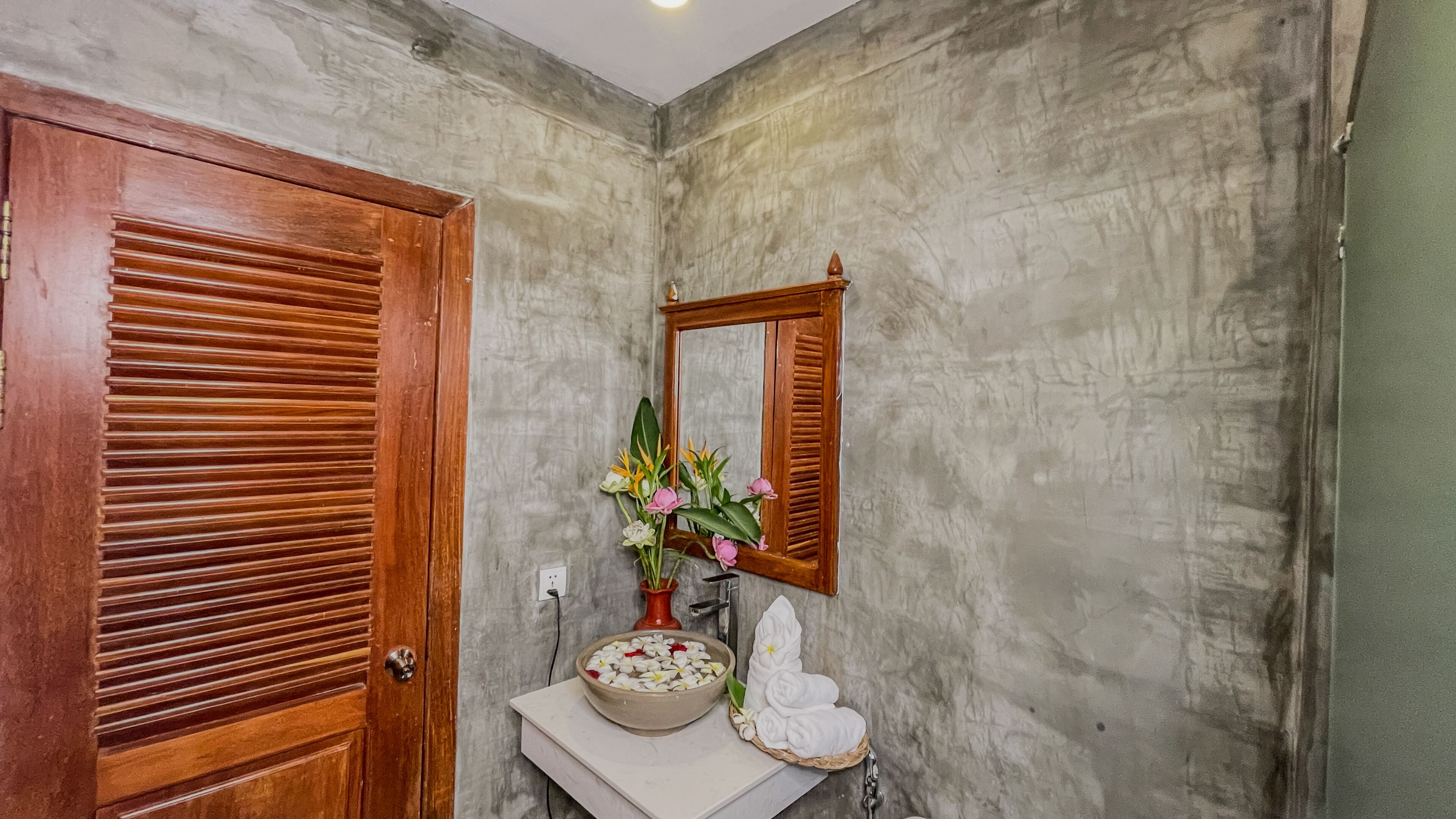 Une salle de bain avec une porte en bois, un miroir, un lavabo agrémenté d’un vase de fleurs et un porte-serviettes.