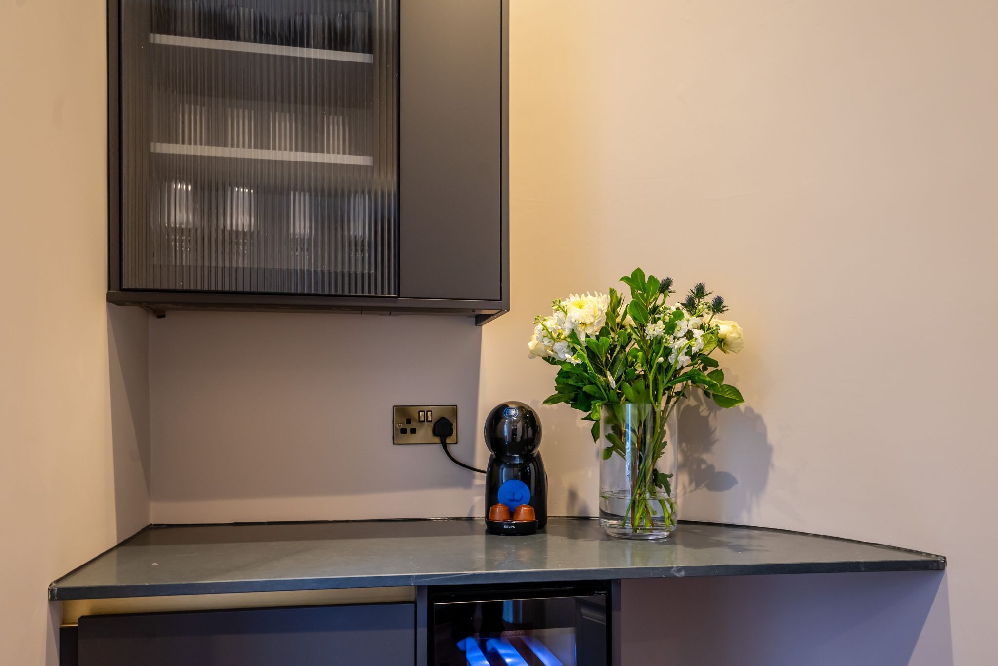 Un comptoir de cuisine avec une machine à café, un vase de fleurs et un meuble suspendu au-dessus.