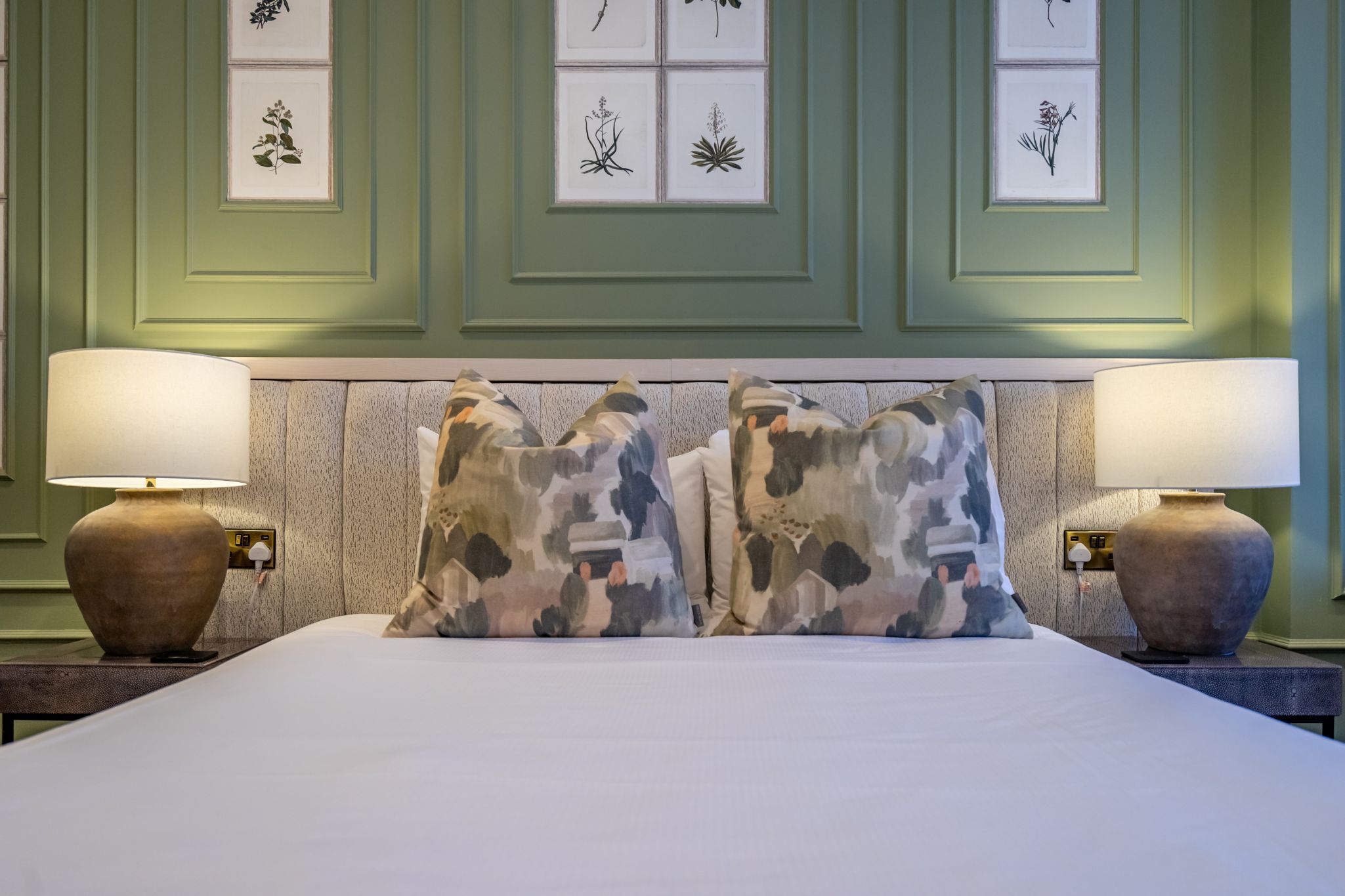 A neatly made bed with two lamps on either side, a headboard with framed botanical prints, and a textured wall panel.