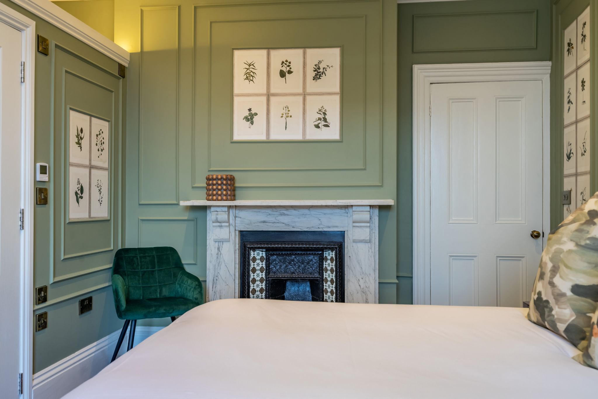 Une chambre à coucher avec un lit, un fauteuil vert, une cheminée et des œuvres d’art botanique encadrées accrochées au mur.