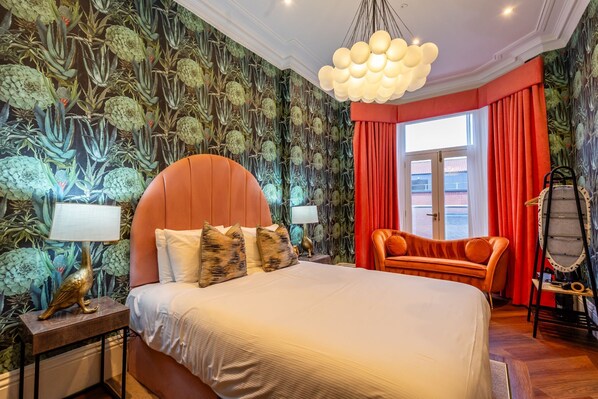 Ein Schlafzimmer mit einem großen Bett, einem rot gepolsterten Sessel, einer Hängeleuchte und einem Fenster mit roten Vorhängen.