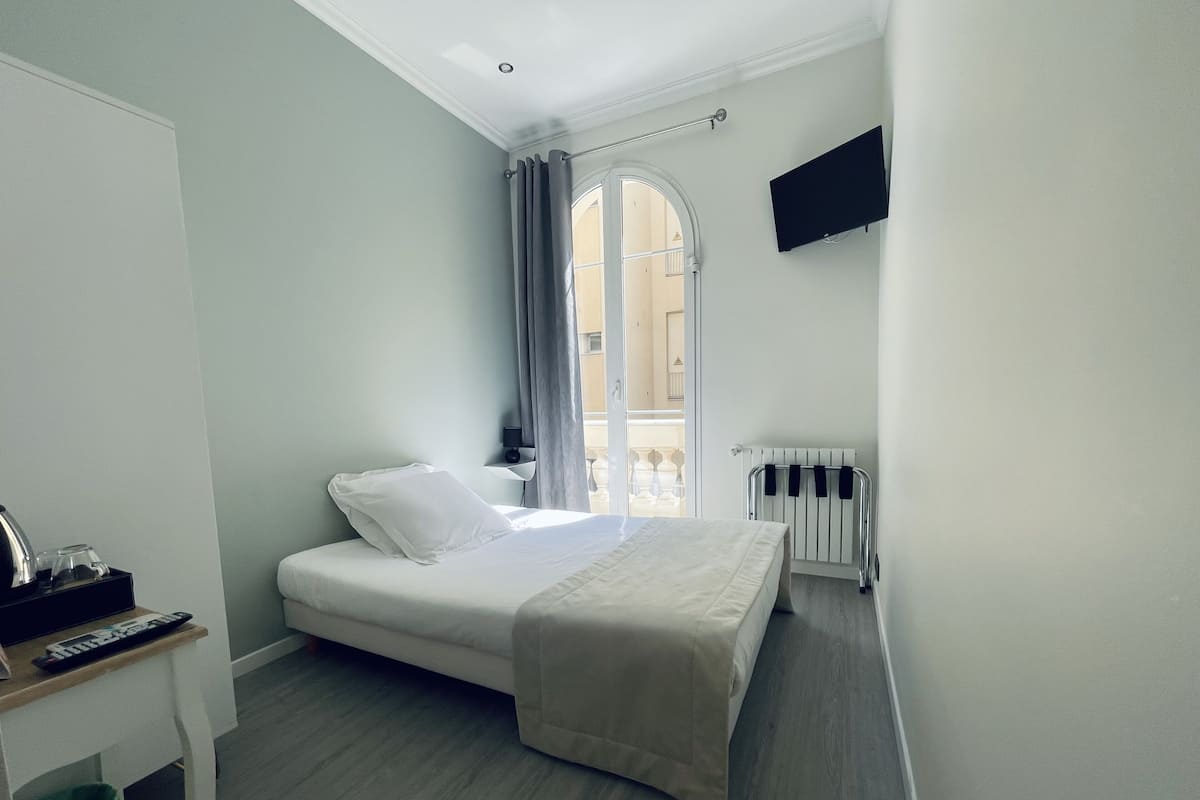 Una habitación de hotel con cama, escritorio con control remoto, televisor montado en la pared y ventana con cortinas.