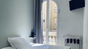 Een keurig opgemaakt bed met witte lakens in een kamer met een witte radiator, een kleine tafel met een lamp en een aan de muur bevestigde televisie.
